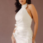 Fabric is Satin, silhouette is Fit and Flare / Modified A-Line, neckline is Halter, sleeve style is Sleeveless, train is Chapel Train, waistline is Natural Ruched, embellishment is 3D Floral Appliqué, back is Low V-Back, color is Ivory, style is Modern Minimalist Romance, Smooth satin with 3D floral details and a removable train for two distinct bridal looks.