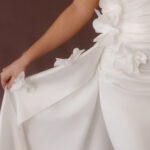 Fabric is Satin, silhouette is Fit and Flare / Modified A-Line, neckline is Halter, sleeve style is Sleeveless, train is Chapel Train, waistline is Natural Ruched, embellishment is 3D Floral Appliqué, back is Low V-Back, color is Ivory, style is Modern Minimalist Romance, Smooth satin with 3D floral details and a removable train for two distinct bridal looks.